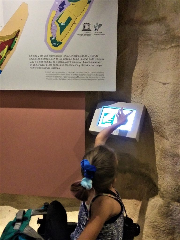 Anna, a teen wheelchair user, is using an interactive touchscreen at the Cozumel Museum in Mexico. She is wearing a blue and white print tank top and has her hair pulled back into 2 blue scrunchies. The walls of this portion of the museum are clay colored or textured to appear like rough rock.