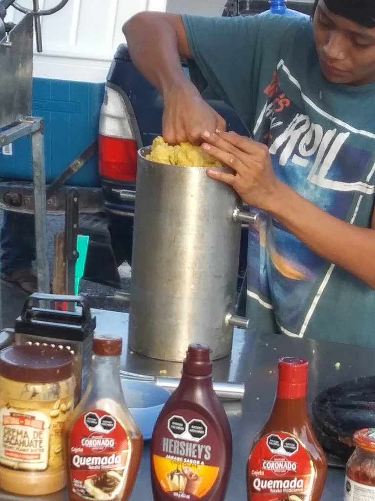 Youth in streetside churro stand using his fist to pack down the dough inside the canister of the machine used to form the churros. Bottles of toppings line the table in front of him.