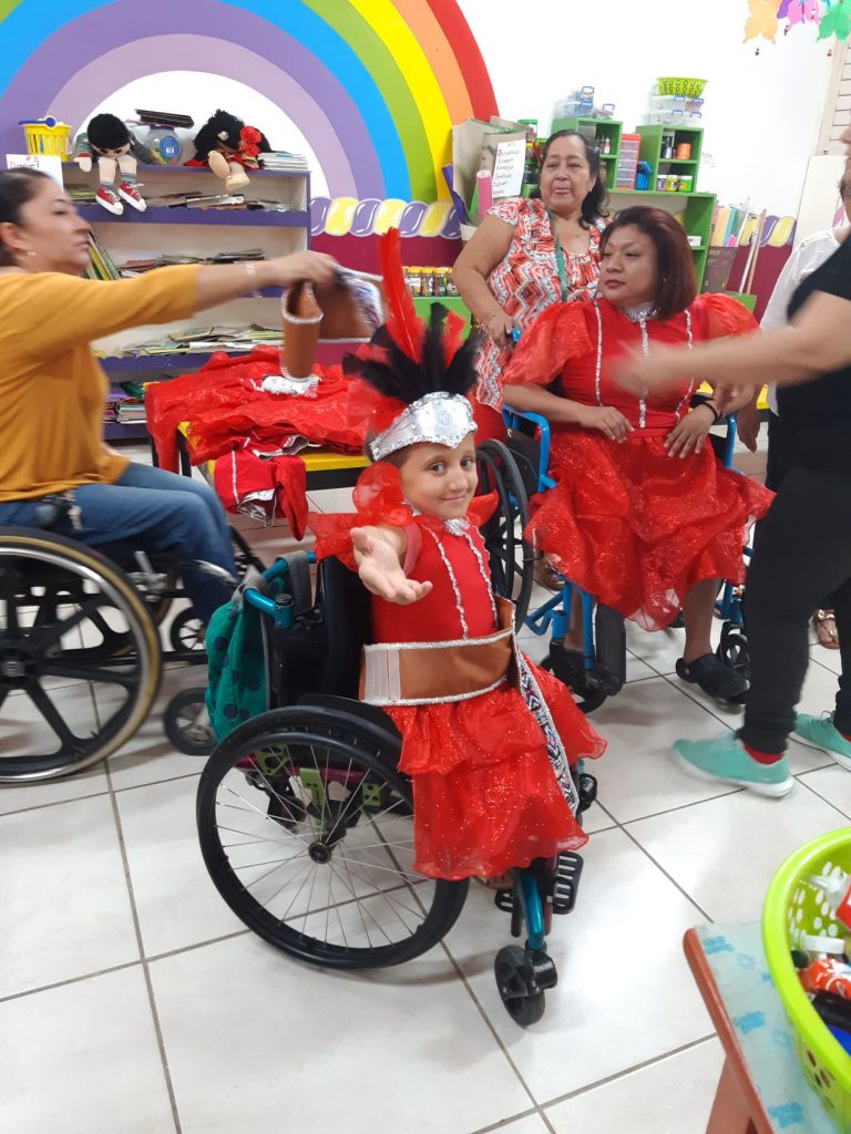 Anna a caucasian teen wheelchair user at a costume fitting for her Cozumel Carnaval dance performance. She is turned posing for the camera with her hand extended, and others are gathered near a table holding costumes.