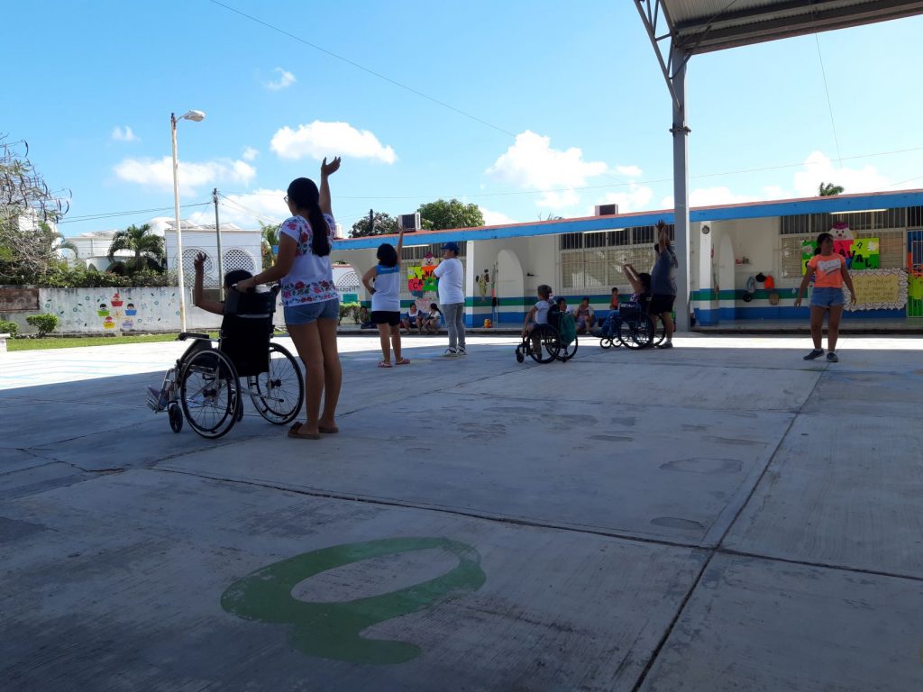 A group of wheelchair users in Cozumel Mexico at a dance practice session in a school playground. Anna is the dancer in the center of the photo. All youth are seen from behind.