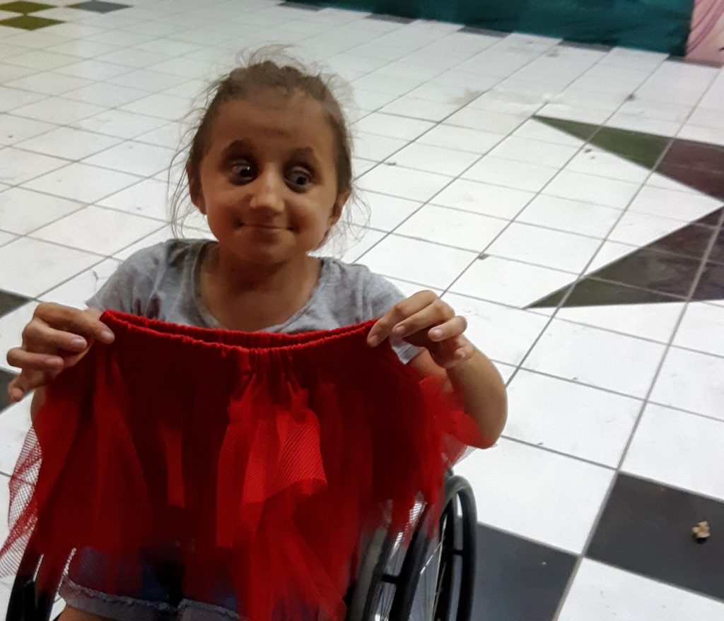 Anna, a caucasian teen wheelchair user, holding up a short fluffy red skirt prototype and looking skeptical.