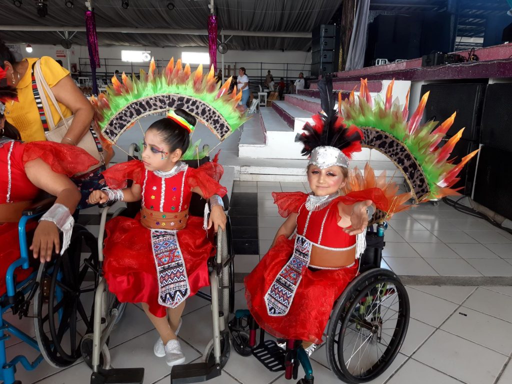 Anna is posing for the camera with her arm outstretched as she sits next to other members of her dance troupe using wheelchairs. They are wearing red dress costumes and the feathered back pieces for their wheelchairs are visible. They are gathered before their performance.