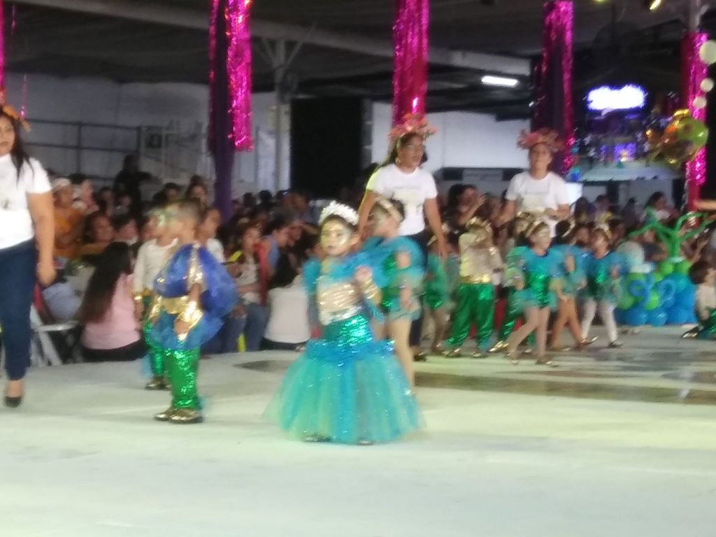 A group of preschool age children in aqua, blue and green costumes perform in a youth dance competition as part of Cozumel Carnaval. Their dance instructors are along the edge of the stage providing supervision and instruction.