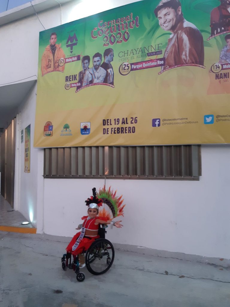 Anna, a teen wheelchair user in her Carnaval costume posing beneath a large advertising banner for the concert series that is part of Carnaval season in Cozumel Mexico.