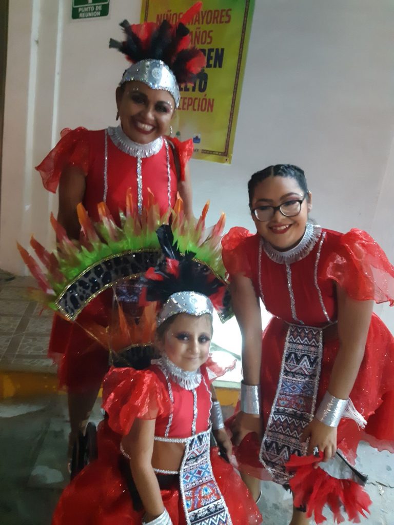 Anna and two other members of her Cozumel Carnaval dance troupe smile and pose. They are wearing red and black feathered headpieces with red costume dresses. Anna is seated in her wheelchair with the large feathered fan shaped backpiece attached.