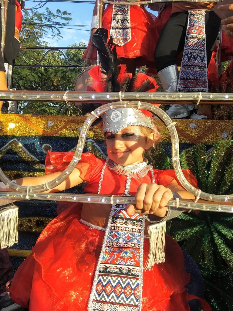 A close-up shot of Anna a teen wheelchair user riding on a Carnaval parade float in Cozumel. She is holding onto a lighted railing and smiling as she looks through the railing. She has her long hair pulled back under the feathered headpiece and is wearing dramatic eye makeup for the performance.