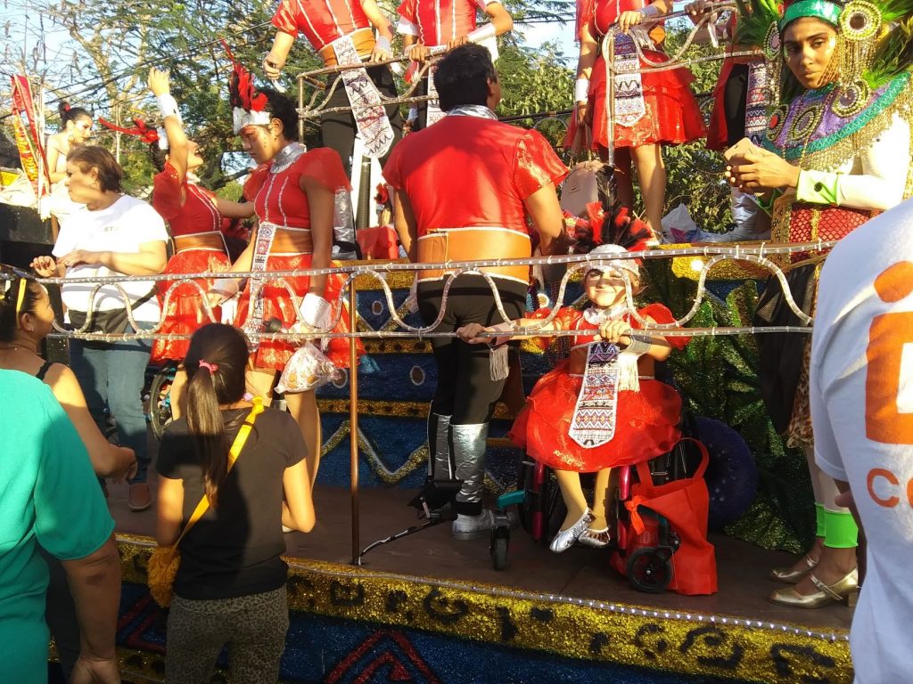 Anna is seated in her wheelchair along the first level of the jaguar themed float. She is holding onto the railing and looking through it. She is wearing a red full skirted costume with a feathered headpiece and smiling. There are other members of her dance troupe on upper levels of the float. They are loading and preparing for the parade launch.
