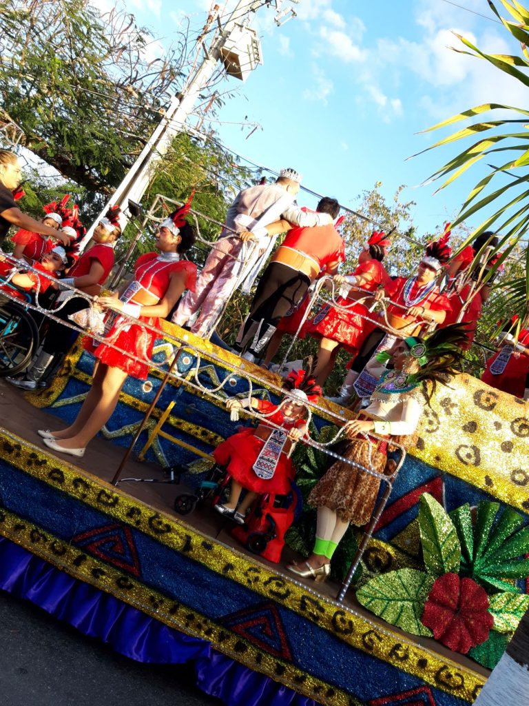 Anna is seated in her wheelchair along the first level of the jaguar themed float. She is holding onto the railing and looking through it. She is wearing a red full skirted costume with a feathered headpiece and smiling. There are other members of her dance troupe on upper levels of the float. They are loading and preparing for the parade launch. This photo is tipped at a 45 degree angle.