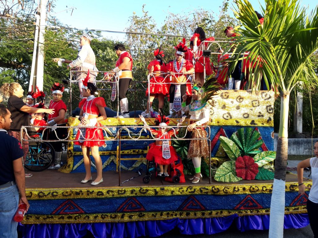 Anna, with her arms raised is seated in her wheelchair along the first level of the jaguar themed float. She is wearing a red full skirted costume with a feathered headpiece and smiling. There are other members of her dance troupe on upper levels of the float. They are loading and preparing for the parade launch.