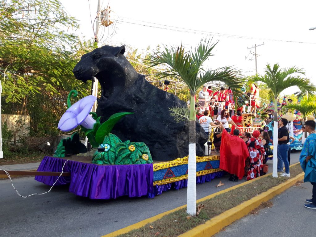 A huge sculpted jaguar with his mouth open wide, is at the front of this Cozumel Carnaval float. There are large tropical flowers and faux animal fur strips embellish the sides of the float. The float is being loaded with youth in red costumes.
