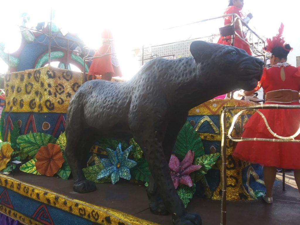 A large sculpted jaguar is part of this Cozumel Carnaval float. There are large tropical flowers and faux animal fur strips embellish the sides of the float. There are several youth in red dress costumes riding onboard the float.