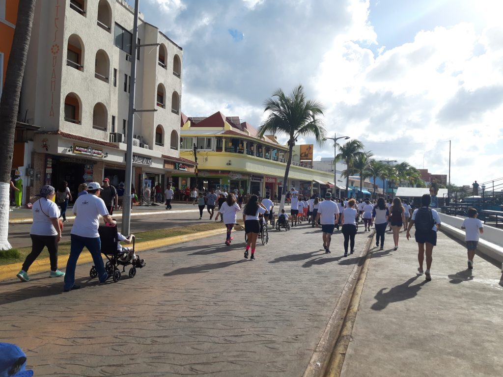 A large group of people have their backs facing the camera as they walk along the Cozumel streets for the Paint and Run. Their white t-shirts are clean and neat. In the distance Anna rolls ahead in her wheelchair.
