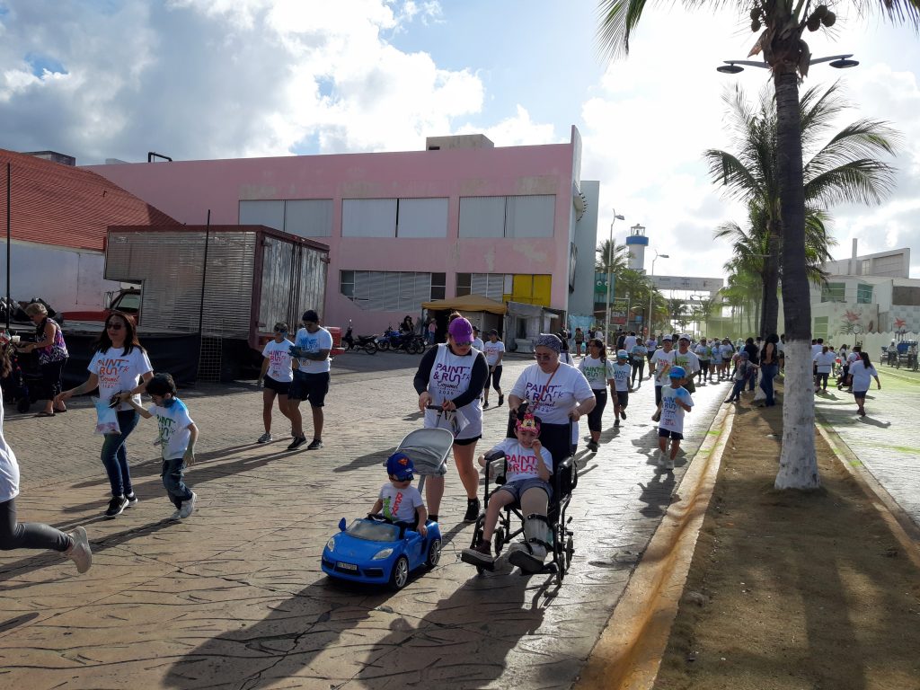 A landscape photo shows a large group of people donning white t-shirts that say Paint and Run. Some push strollers while one person pushes their friend in a wheelchair. Anna is not in this photo.