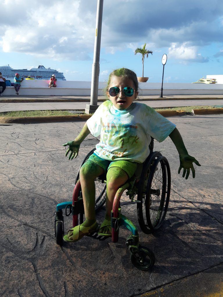 A caucasian teenage girl in a wheelchair strikes a pose, pretending to be shocked at how messy she has gotten from all the green and blue paint that was thrown during the Paint and Run.