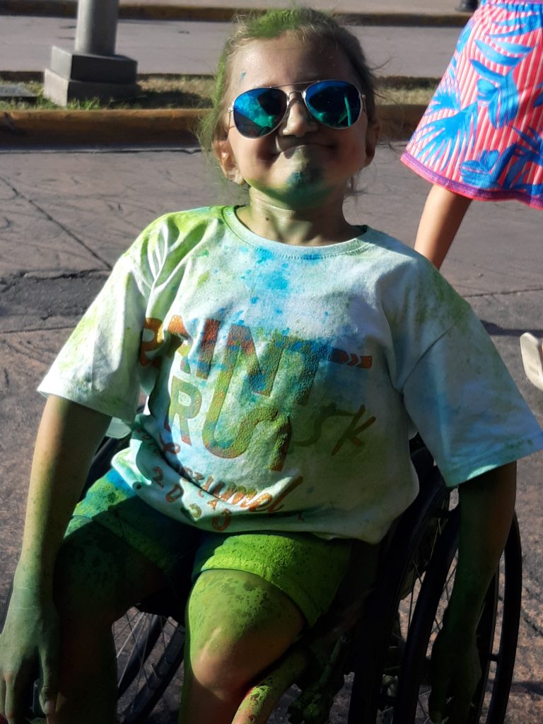 Anna, a caucasian wheelchair-using teenager smiles at the camera. Her skin is stained with blue and green paint, as well as her Aviator glasses.