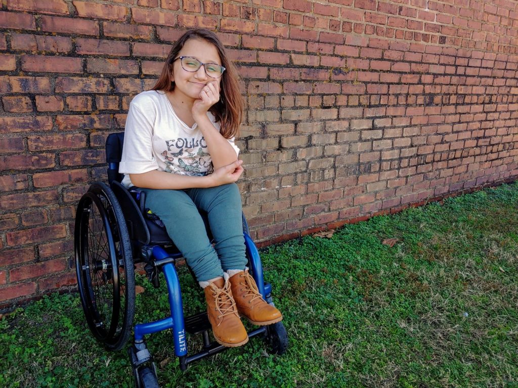 Teen girl poses in her a sporty, navy blue wheelchair. She's wearing a cream colored shirt with butterflies on it, leggings, and cute brown boots. She is posing in front of an old brick wall with grass below her.