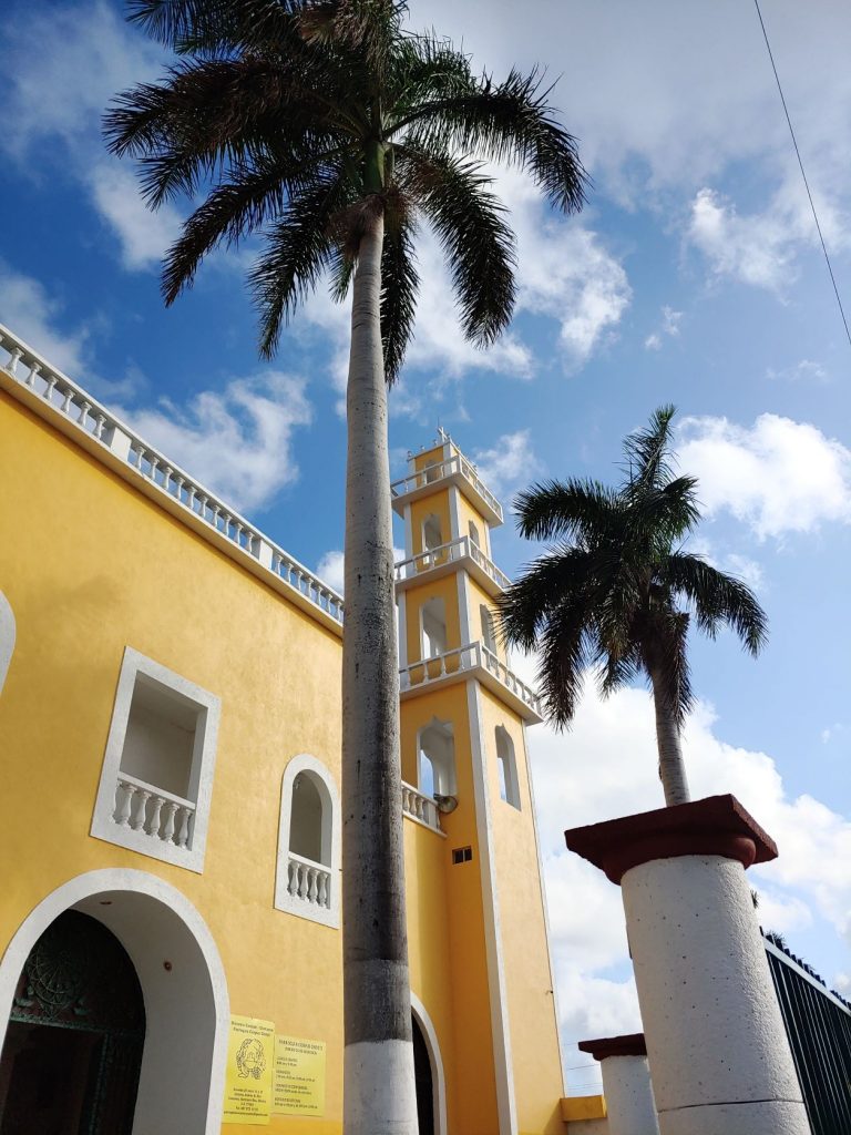 A view of the vivid yellow with white trimmed Corpus Christ Cathedral in San Miguel, Cozumel, Mexico. An arched entry is shown along with a tower and two tall palm trees. There are a couple old freestanding pillars in the foreground. Blue cloudy sky above.