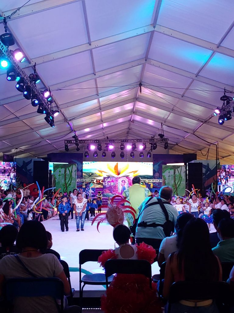 A dance performance with children for Carnaval. The children are in matching costumes and their mothers hold their hands and guide them through the performance. The mothers are wearing matching t-shirts and jeans.