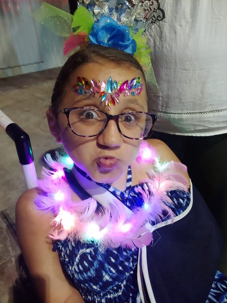 A teen girl wearing a blue dress with gems on her forhead sits in a wheelchair about to enjoy the parades of Cozumel's carnaval celebration showing the accessibility.
