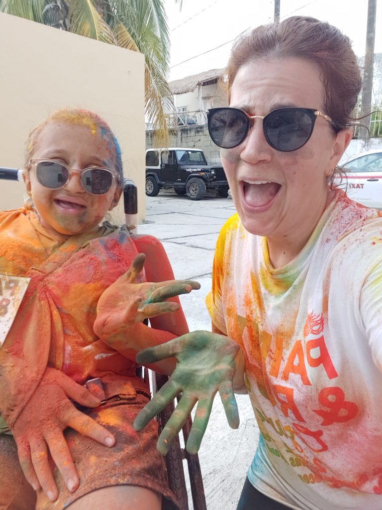 Anna and her mom make silly expressions with their paint-covered faces. Anna uses her uninjured arm to raise it as part of her funny pose. Her wheelchair wheel is stained with paint.