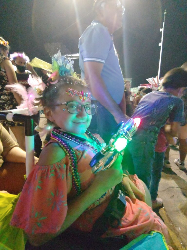 A teen girl poses in her wheelchair with a light up toy laser gun while along a dark street awaiting a Cozumel Carnaval parade.