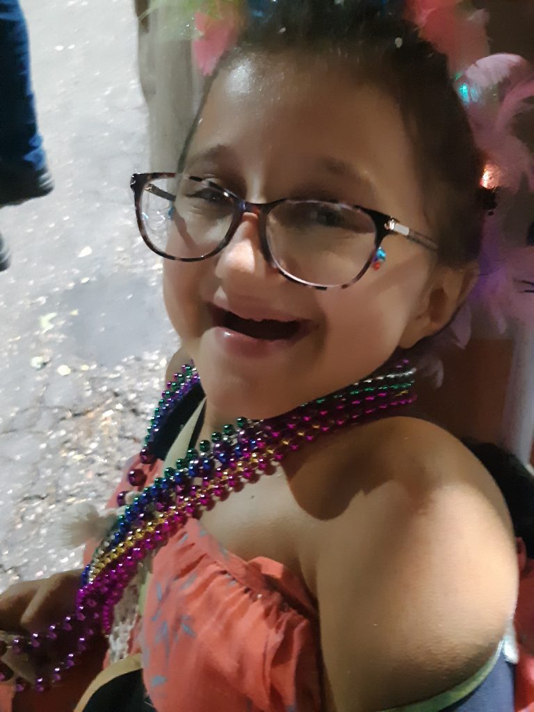 A teen girl wearing a pink dress and beaded necklaces sits in a wheelchair about to enjoy the parades of Cozumel's carnaval celebration showing the accessibility.
