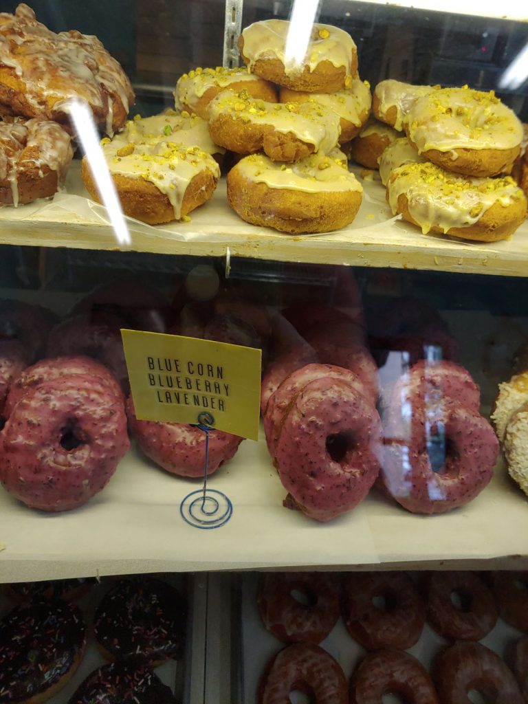 A view into a glass front bakery case showing an upper shelf with yellow frosted donuts topped with fresh lemon zest and a lower shelf with Blue Corn Blueberry Lavender donuts. They appear purple-ish and have a light gritty glaze.