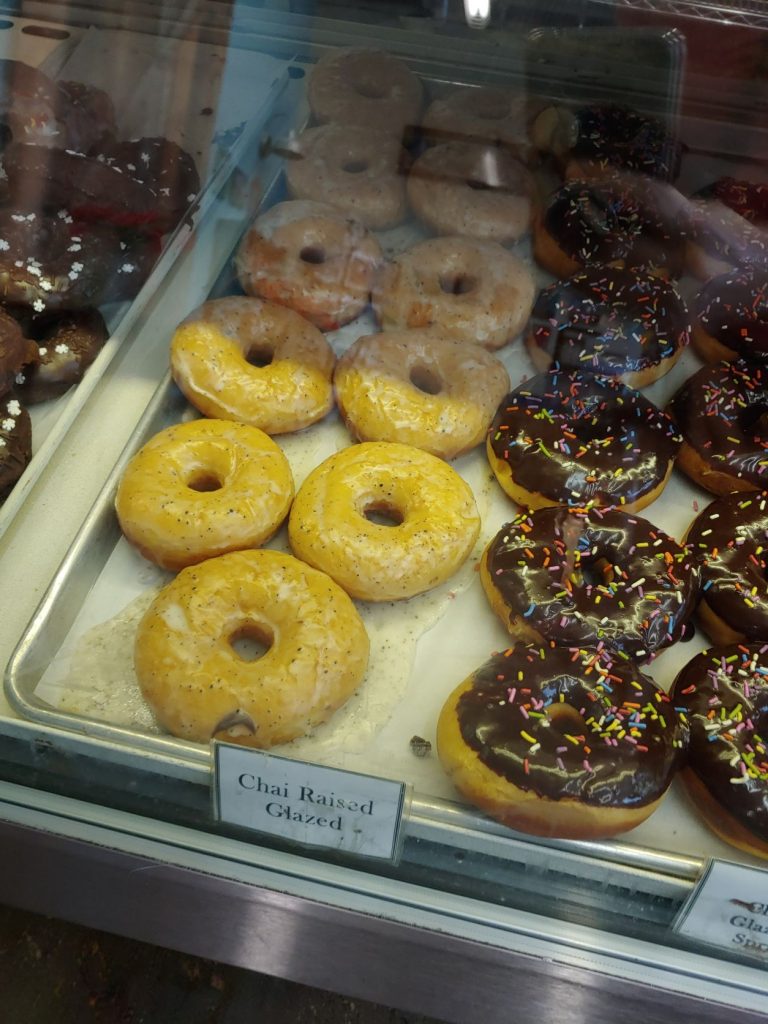 A view into a glass bakery case at Whoo's Donuts with a parchment paper lined metal bakers tray holding Chair Raised Glazed and Chocolate Frosted with Sprinkles donuts. There is a small sign indicating the flavors in front of each style.