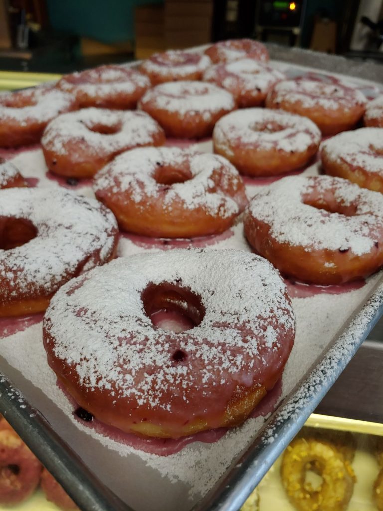 A metal bakers tray lined with parchment paper with rows of hot donuts topped with sprinkled powdered sugar.