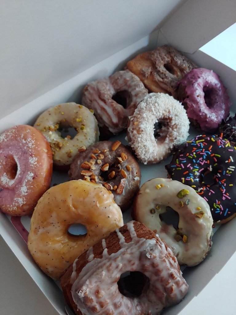 An open white donut box with a dozen mixed donuts inside. There is a broad variety with chocolate or vanilla frosted, glazed and creamy iced styles topped with nuts, sprinkles, coconut and sugars.