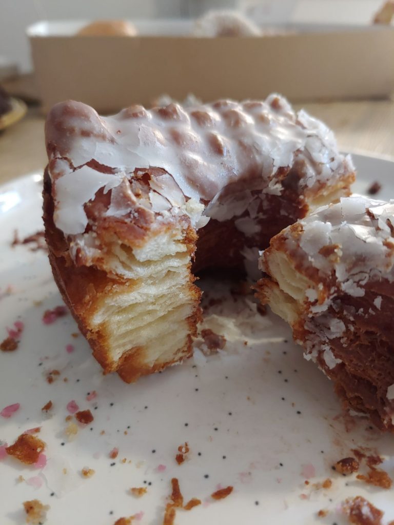 A cro-nut cut open to show all the layers of pastry inside. It has a white icing and is on a white plate with crumbs.