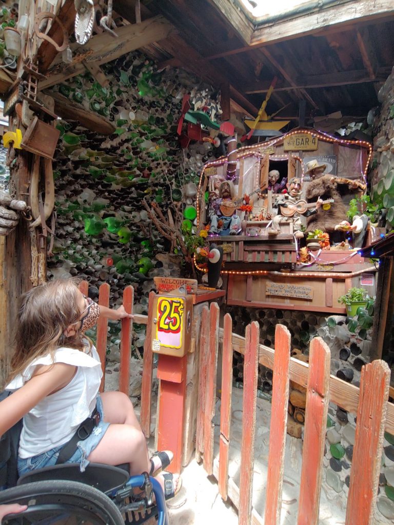 Anna, a teen girl in denim shorts and a ruffled white t-shirt, in her wheelchair in front of an interactive mechanical diorama of a country bar band. There is a brown wooden fence surrounding the scene and a sign indicating 25 cents. She is looking at the band.