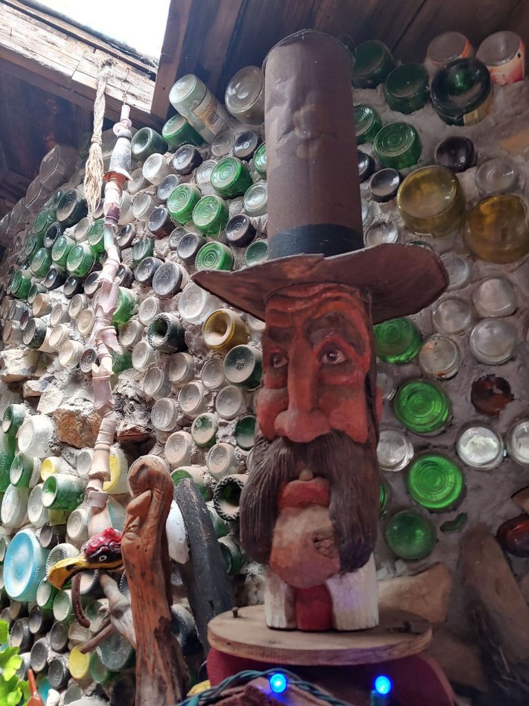 A tall wall built from old glass bottles, mostly clear or green,, stacked on top of each other and cemented together. There is a large folk art sculpture of a man with a long mustache wearing a very tall top hat. There is light above coming from a skylight.