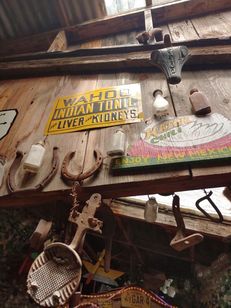 View of relics and old advertising signs hanging high on a wood wall and from rafters. There are rusty horseshoes, old bottles, old tools. The yellow and black advertising sign reads Wahoo Indian Tonic for Liver and Kidneys.
