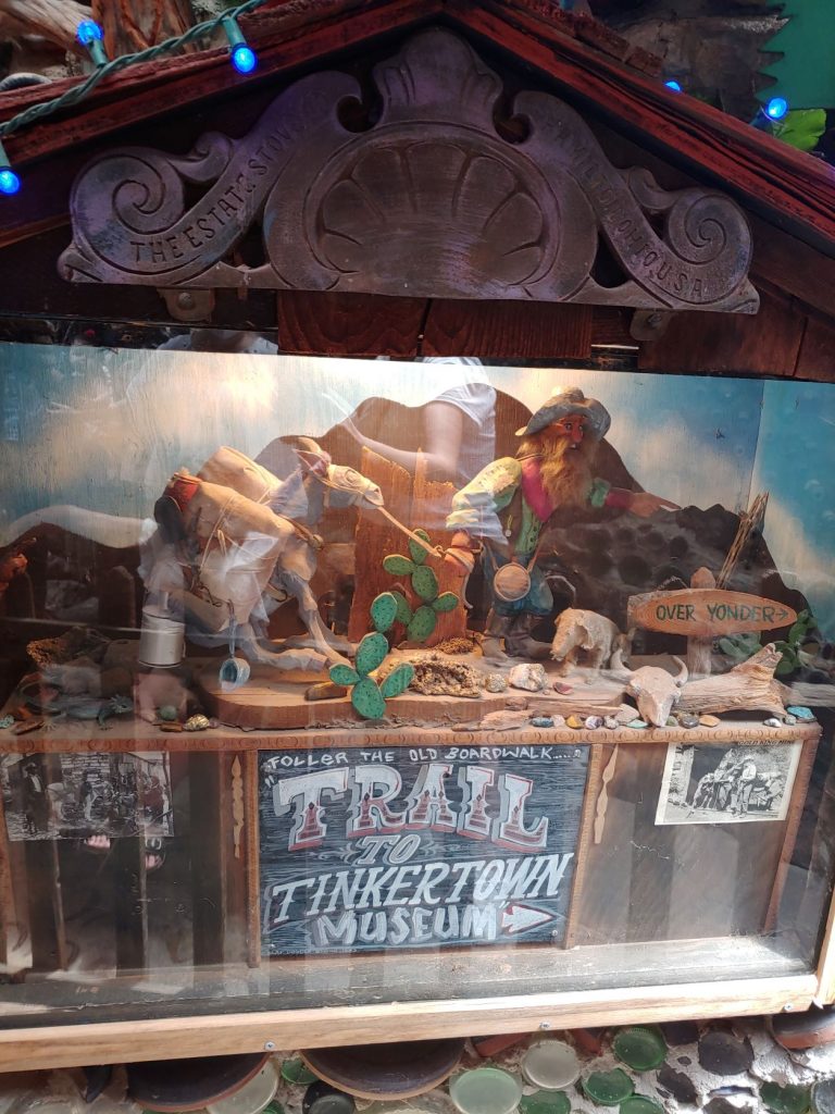 A glass enclosed diorama that has a wild west rocky and hilly landscape with a prospecting scene. Possibly a reference to the turquoise mined locally in the past. A sign below the display appears like a chalkboard with hand lettering. Trail to Tinkertown museum. There are small blue light bulbs above.
