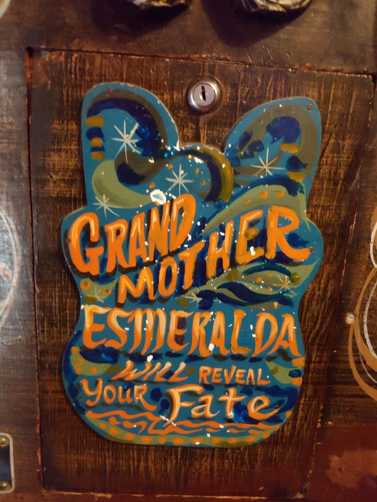 A painted sign on a locking wooden. panel of the fortune teller machine. Dark brown wood with blue and green patterned sign with gold lettering Grand Mother Esmeralda Will Reveal Your Fate.