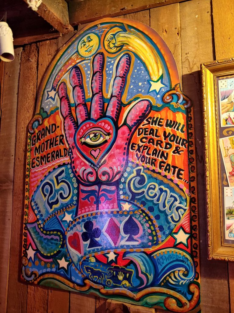 Hand-painted arched wooden sign with an upright pink hand in the center. The palm has a eye in the center of a heart. Grandmother Esmeralda She will deal your card and explain your fate. 25 cents. There are swirls, playing card suits and a sun, moon and stars painted on the sign. Blue and orange background.