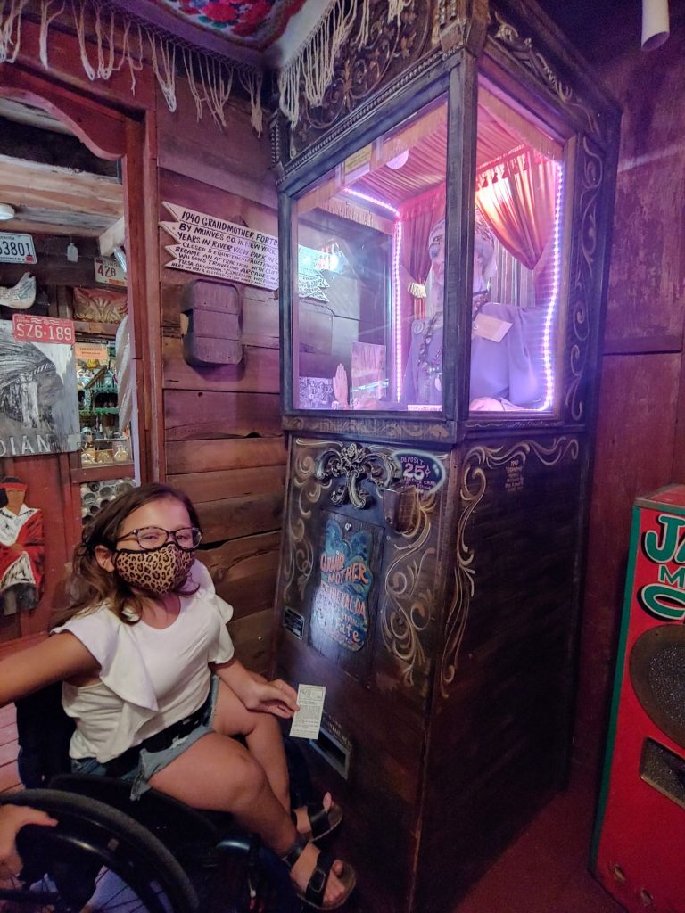 Anna, a teen wearing denim shorts and a white ruffled t-shirt, sitting in her wheelchair in front of the vintage lighted fortune teller machine. She is holding the printed ticket that was dispensed with her fortune. The machine has an ornate black and gold cabinet with an upper window that holds a bust of a gypsy fortune teller.