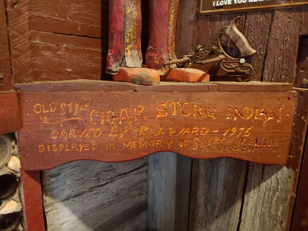 Carved wood sign on a shelf that readsOld Style Cigar Store Indian Carved by R.J.Ward- 1976