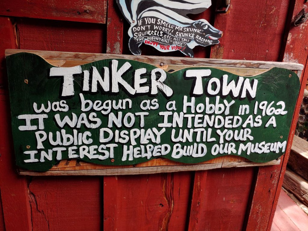 Carved wooden sign with white raised letters on an old red painted wooden door. Sign reads: Tinker Town was begun as a hobby in 1962. It was not intended as a public display until your interest helped build our museum. There is a carved skunk above the sign that says Skunks, racoons,, squirrels, etc. all call the mountains their home.