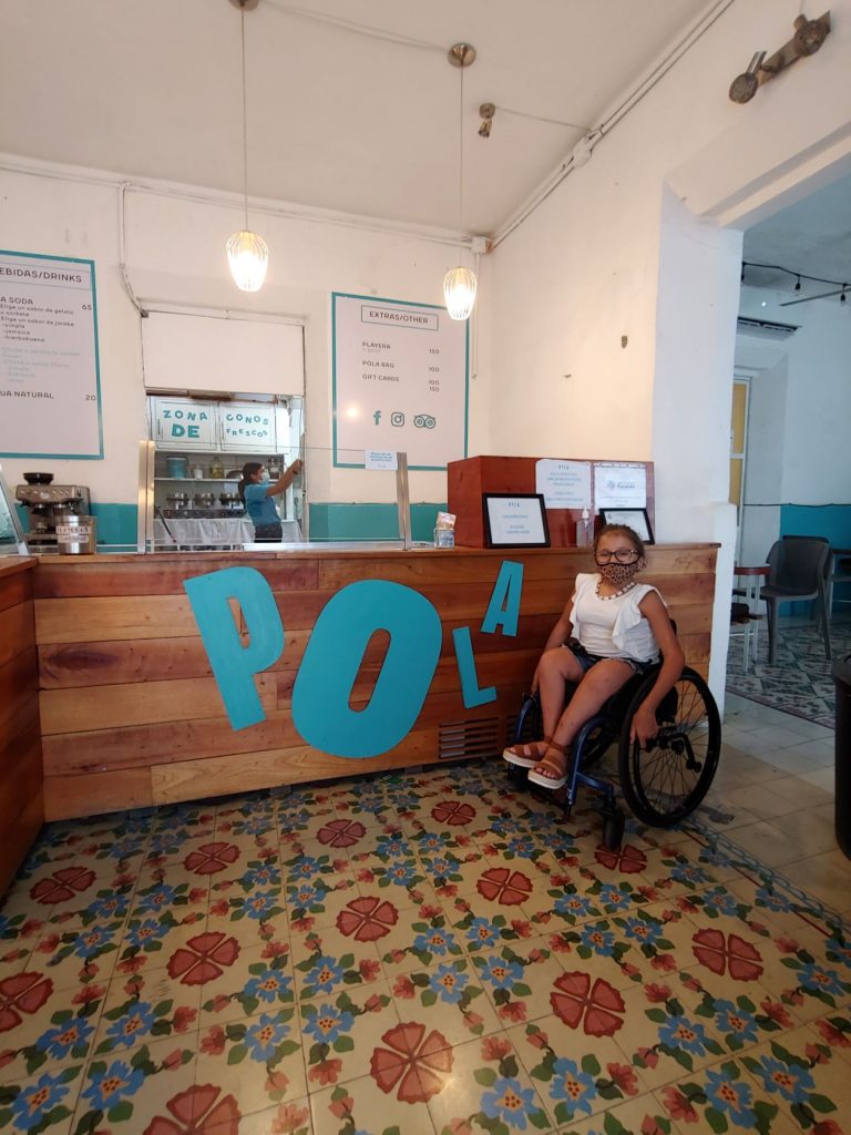 Teen in wheelchair at Pola gelato shop giving accessibility review. There is a multicolored "pasta" tiled floor in front of her. The counter is at her chin height and the front is wood strips with attached turquoise Pola letters.
