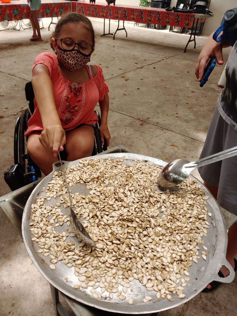 Teen girl in wheelchair roasts pumpkin seeds over an outdoor stove as part of immersive cooking lesson while attending accessible Spanish lessons in Merida, Mexico.
