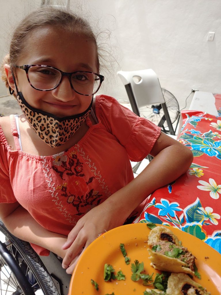 Teen girl using a wheelchair wearing a pink shirt smiles at the camera with the tacos she learned to make during an immersive Spanish Lesson.