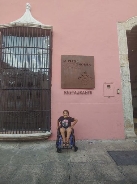 Girl in wheelchair poses in front of restaurant, showing accessible Yucatan dining in Merida.