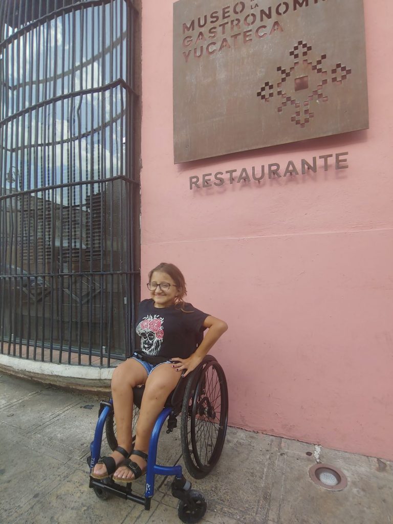 Girl in wheelchair poses in front of restaurant, showing accessible Yucatan dining in Merida.