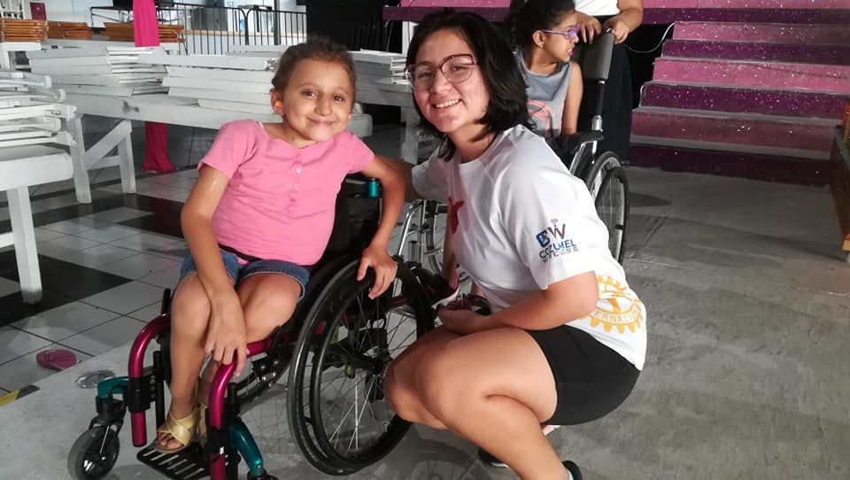 A candid pose of Anna with one of the Carnaval queens during a practice session. Anna is sitting in her wheelchair in a pink t-shirt and jean shorts and the queen is wearing a printed white t-shirt over black athletic shorts and is kneeling down next to Anna.