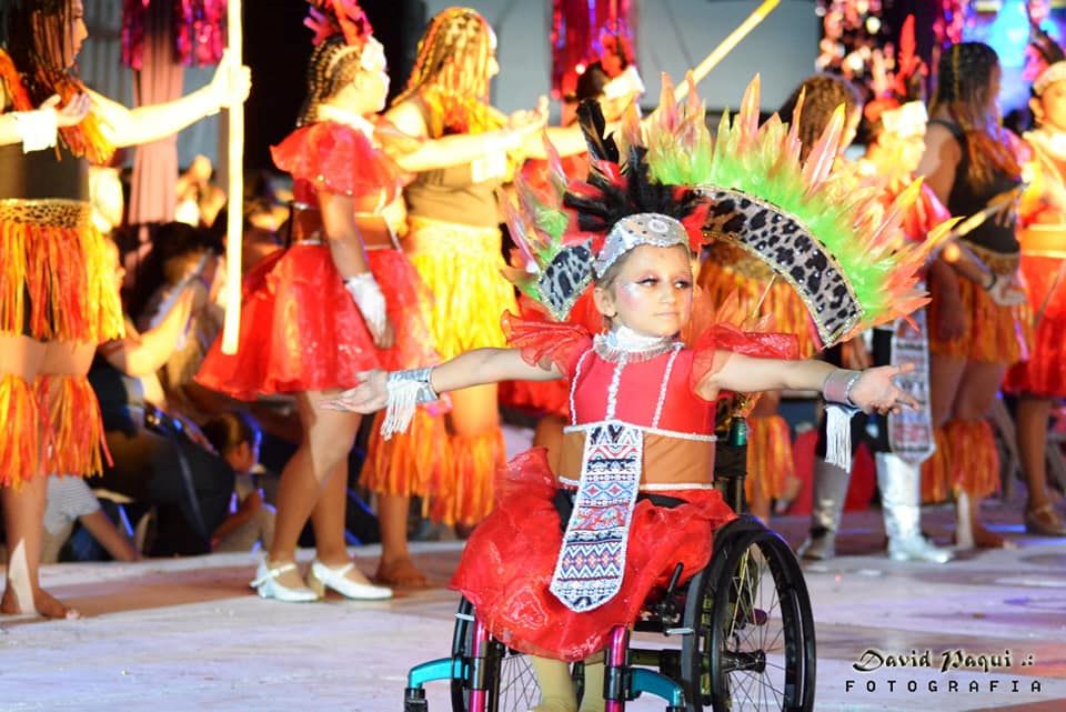 Anna in her wheelchair with her arms extended wide. She is participating in a dance program as part of Cozumel Carnaval. She is wearing a red dress with a full skirt, a tan waist piece, silver fringed forearm covers and a red and black feathered headpiece. Her wheelchair has a large feathered fan shaped backpiece attached to it that extends beyond her. Members of the dance troupe are visible around and behind her. She is wearing glittery stage makeup with dramatic eyes.