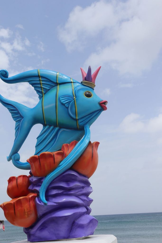 A bright blue angelfish rests on a purple and orange coral base. This is one of the large sculptures that decorate the seawall and malecon of Cozumel during the Carnaval season. A calm ocean and cloudy sky are in the background. The fish sculture is wearing a pink and purple costume hat.