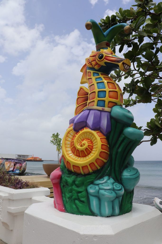Six foot tall whimsical colorful sea horse sculpture for Carnaval season on Cozumel. The seahorse is shown on a pedestal of coral and wearing a Carnaval costume with a jester style hat. The colors are purple, green and gold with a bit of bright pink. The sculpture rests on a white cement seawall and the ocean with a ferry is visible in the background.