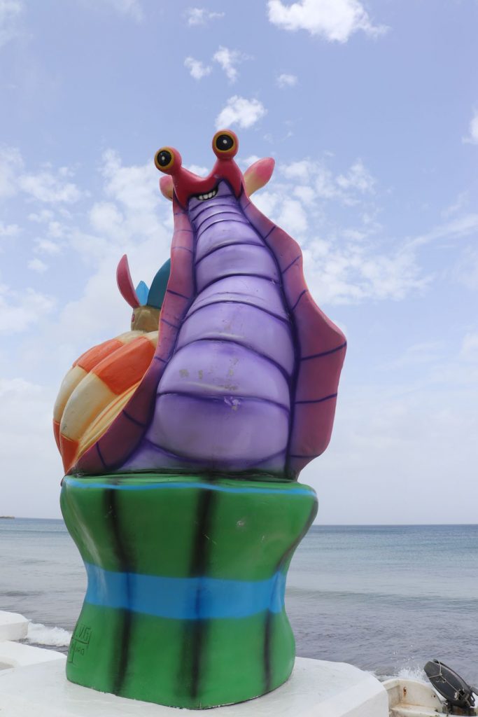 A brightly colored whimsical sea snail sculpture rests on a white cement pedestal along the seawall on the east side of Cozumel. The decoration is green, purple and gold and is maintained through the Carnaval season. They are about 6 feet tall after the base.
